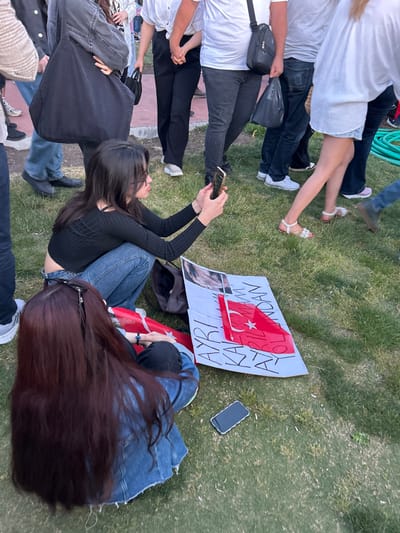 The image shows a scene in a park in İzmir, Türkiye, near the coordinates 38.437, 27.141. It is a sunny day with green grass, and a group of people are gathered around a sign. The sign is white with black text and depicts a red Turkish flag. A young woman with long dark hair is holding a black smartphone in her right hand and taking a picture of the sign. She is wearing a black long-sleeved shirt, a dark blue denim skirt, and jeans. A young woman with long dark brown hair is sitting on the ground in front of the sign, wearing a blue denim jacket and dark blue jeans. She is looking down at her phone, which is laying on the grass. The woman in the background is wearing a white long-sleeved shirt and dark jeans and a person is standing behind her. The woman is using a camera phone. The sign reads "AYRI KARIŞMAM ZORLUNDAN..." with a red Turkish flag. The grass is green and lush. There are other people in the background, not visible in the image, likely attending an event. The general atmosphere is relaxed and casual, reflecting the enjoyment of the public gathering.
