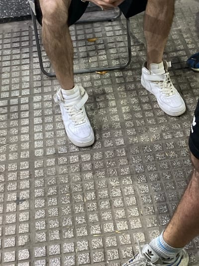 The image depicts an outdoor ground-level view in San Antonio de Padua, Argentina. The scene focuses primarily on the lower extremities of two individuals seated on a grey, square-patterned paved surface composed of exposed aggregate tiles.

The central figure, positioned slightly to the left, is a male with visible bare, hairy legs wearing dark shorts. This individual is seated on a dark, thin-framed metal chair, with their feet clad in white high-top sneakers featuring a prominent ankle strap and white laces. The hands, partially visible at the top of the frame, suggest the person is holding or interacting with a mobile phone.

To the lower right, the bare, hairy leg of a second male individual is visible. This person is wearing black shorts, a grey athletic shoe with visible Adidas branding, and blue and white striped socks.

The paved ground surface is generally grey with some visible dirt and scattered dry, light-colored leaf debris. The lighting is even, suggesting an outdoor environment during daytime or under bright artificial light. No explicit interactions between individuals are visible.