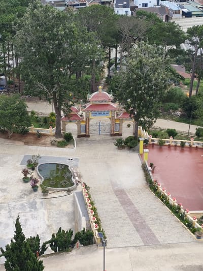 The image captures an aerial view of a temple complex in Lộc Quý, Vietnam. The central focus is a traditional temple gate with a red-tiled roof, adorned with ornate yellow and blue decorations. Beyond the gate, a courtyard is paved with patterned tiles, and a large, red-tiled area is visible to the right, likely an outdoor space for gatherings or ceremonies.
To the left of the gate, a low, ornate yellow fence encloses a garden area with trees and bushes. In the foreground, a heart-shaped pond filled with green water and lotus plants adds a serene element to the scene. Various potted plants are strategically placed throughout the complex, enhancing the greenery. A tall pine tree stands prominently near the gate, casting a shadow over the paved courtyard.
The surrounding environment is characterized by lush vegetation, including numerous trees and dense foliage, suggesting a semi-rural or well-landscaped urban setting. Residential buildings and greenhouses are visible in the background, indicating the presence of a community nearby. The overall atmosphere appears peaceful and well-maintained, typical of a religious or cultural site. There are no people visible in the image.
