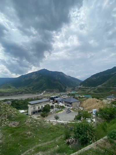 A wide-angle, high-angle shot captures a village nestled in a valley between large, verdant mountains under a cloudy sky. In the foreground, a grassy, uneven slope leads down to a collection of buildings. Several multi-story structures are under construction, with exposed concrete and scaffolding visible. One completed building has a dark, pitched roof and appears to be a residential or commercial property. A gravel parking lot next to it holds a single silver SUV. Further into the scene, more buildings with various roof colors are visible, along with trees and a dusty, excavated mound of earth. In the middle ground, a wide, dry riverbed cuts through the valley, with a distant dam or structure visible on the right. The background is dominated by the imposing, green, and rocky mountains that frame the valley. The sky is overcast with dense, gray clouds, suggesting a temperate or cool climate. The overall scene depicts a developing settlement in a mountainous, rural landscape, identified as the city of Chalda, Russia.