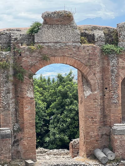 The image depicts a view through an ancient Roman archway constructed of weathered red bricks and stone, possibly part of the ancient Roman theater in Taormina, Sicily, Italy. The arch frames a lush green forest canopy and a distant, hazy landscape with mountains in the background, one of which appears to be Mount Etna. The stonework of the archway shows signs of age and repair, with vegetation growing in crevices. In the foreground, there are scattered stones and two cylindrical stone remnants lying horizontally. The sky above is cloudy. No people are visible in the scene.