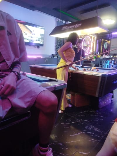 The image depicts a dimly lit billiards hall, likely in Patong, Thailand, with a focus on a woman in a yellow dress playing pool. She is positioned near the middle of a pool table, holding a cue stick and preparing to strike a ball. The table itself is well-lit by overhead lamps, revealing a scattering of billiard balls. To the left, a person is partially visible, seated and wearing a light-colored uniform or shirt, with visible arm tattoos and a bracelet. In the background, another person, a man, stands behind the pool table, also holding a cue stick. Shelves stocked with bottles are visible further in the background, suggesting a bar area. The floor is a dark, marbled tile. The overall atmosphere is that of a recreational establishment, with neon lighting and a television screen displaying an image in the upper left corner.