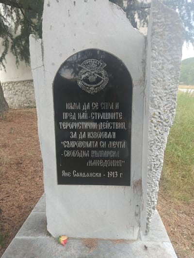 This media file depicts a gravestone or memorial monument outdoors. The monument is made of stone, with a black plaque inset into a larger, rough-hewn white stone slab. The black plaque contains text in white Cyrillic script and a circular emblem at the top. The emblem features a flag with the words "СВОБОДА ИЛИ СМЪРТЬ" (Freedom or Death) and an image of a hand holding a bomb and a rifle. Below this, more text reads "ЗА ПОРОБЕНАТА РОДИНА МАКЕДОНИЯ" (For the enslaved motherland Macedonia).
The main inscription on the plaque, attributed to "Яне Сандански - 1913 г." (Yane Sandanski - 1913), speaks of not stopping before the most terrible terrorist actions to achieve the dream of a "free Bulgarian Macedonia."
The monument is situated outdoors, likely in a cemetery or historical site. The background shows dry grass and some trees, suggesting a rural or natural setting. A small, wilted rose lies at the base of the monument. The lighting and scene suggest it is daytime. The overall tone is somber and commemorative.
