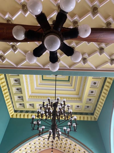 This image captures a detailed, upward-looking perspective of an ornate interior space, likely a grand hall or public building in Pernik, Bulgaria.
The left side of the frame features a distinctive lighting fixture centered around a polished wooden propeller blade. Attached to the hub and extending outwards are multiple arms with frosted white, spherical light bulbs. The word "Heine" is visibly inscribed in a cursive font on the wooden blade. The ceiling behind this fixture is light yellow, adorned with a prominent white zigzag pattern and interspersed with small, gold-colored floral motifs.
The right side showcases a large, elaborate chandelier crafted from dark metal, featuring numerous frosted white light bulbs arranged radially around a central axis, resembling a spoked wheel. The ceiling and wall structures on this side are extensively decorated with multiple layers of architectural moldings and recessed panels. These elements display a vibrant palette of light yellow, white, and teal, with several panels containing small, gold-colored square motifs with floral centers. Further to the right, a large curved ceiling section transitions from teal to an arched area featuring a yellow diamond grid pattern.
The overall aesthetic is one of grandeur and intricate detail, characterized by the dominant colors of light yellow, teal, white, and gold accents. The focus is entirely on the decorative architecture and lighting, with no people or specific activities depicted.