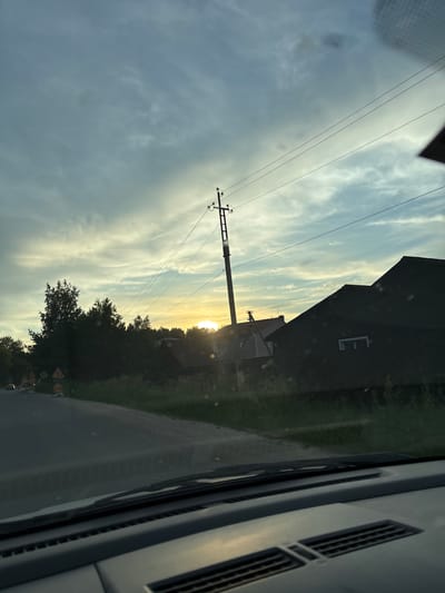 The image depicts a rural roadside scene with a dramatic sky at sunset, viewed from inside a vehicle. The perspective suggests the camera is positioned on the dashboard, with the windshield in the foreground. The sky is a blend of blues, grays, and yellows, with wispy clouds streaked across it. The setting sun is a bright, hazy orb peeking through the clouds, casting a warm glow on the scene.
On the right side of the image, a dark, angular house with a prominent pitched roof dominates the middle ground. A single, rectangular white window is visible on its dark facade. To the left of this house, a utility pole with multiple wires stretches vertically into the sky, bisecting the frame. Further left, a dense line of trees forms a dark silhouette against the brightening sky.
In the foreground on the left, a paved road curves gently to the left, disappearing into the distance. Several traffic signs are visible along the side of the road, including caution signs with yellow triangular shapes. A single vehicle is partially visible on the road in the far distance. The grassy verge beside the road is overgrown with weeds and wildflowers. The overall impression is of a quiet, possibly remote, location bathed in the soft, diffused light of dusk. The scene is devoid of any identifiable human figures. The location is identified as Chigiri, Russia.