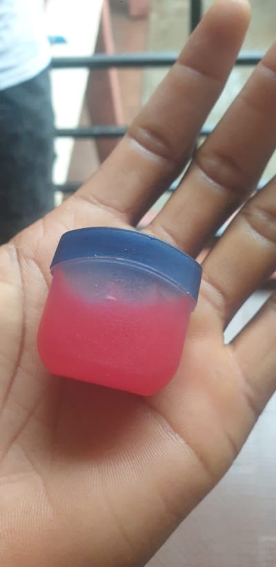 In Yelwa, Nigeria, a person's open hand, likely held in a slight cupping gesture, supports a small, square, translucent plastic container with a dark blue lid. The container is filled with a vibrant pink substance, possibly a lip balm or a similar cosmetic product, which appears somewhat granular. The skin of the hand is dark-skinned, with visible fingerprints and creases on the palm and fingers. In the blurred background, a metallic railing and indistinct figures can be observed, suggesting an outdoor or semi-enclosed urban setting.