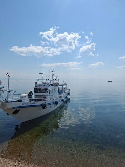 A white passenger boat named "ТЕНГРИ" (Tengri) is docked on the clear, shallow waters of Lake Baikal in Listvyanka, Russia. The boat, bearing the registration number "ВС-02-116", is white with blue trim and features black fenders along its side. A person wearing a hat is visible on the deck, leaning against a railing. In the background, the calm water stretches to the horizon under a bright blue sky dotted with fluffy white clouds. Another boat is visible in the distance. The shoreline in the foreground is covered with small pebbles. The water is so clear that the rocky bottom is visible beneath the surface.