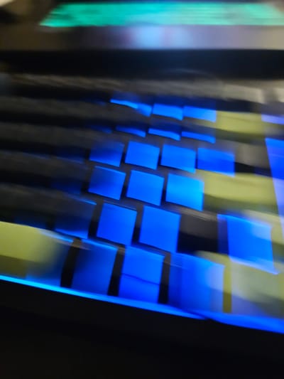 The image shows a close-up, blurred, abstract view of a keyboard with illuminated blue keys and a faint green light at the top. The perspective is angled, and the motion blur suggests movement or a long exposure. The scene is dark overall, with the blue light from the keys being the most prominent feature. There is no visible indication of the city of Versailles, France, or any specific geographical location. The image is purely an abstract visual representation of a keyboard.