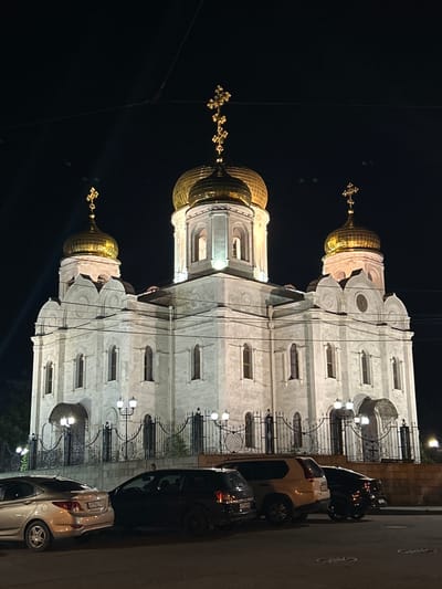 The image depicts a view of a grand, ornate building, likely a church, with golden domes topped by crosses. The building's facade is constructed of light-colored stone with numerous arched windows and decorative architectural elements. The scene is illuminated at night, casting a warm glow on the building and the street. Several cars are parked along the side of the road in front of the building, including a silver hatchback and a darker sedan. The street itself is paved and appears to be in an urban setting. The overall atmosphere is one of nighttime tranquility and architectural grandeur. The image is taken from a slightly angled perspective, with the building dominating the frame. The time of day is clearly night, with a dark sky serving as the backdrop. There are no visible people in the image. The location is hinted to be Pyatigorsk, Russia.