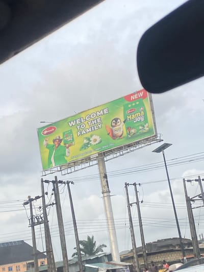 A wide-angle shot captures a billboard advertising "Mama Joy" detergent, positioned above a street scene in Shangisha, Nigeria. The billboard, tilted and angled upwards, features a smiling woman in a green outfit and a hat, holding a green packet of Mama Joy. To her right, a penguin character wearing a shirt with "Mama Joy" printed on it, holding a surfboard, is depicted. The billboard's text reads "WELCOME TO THE FAMILY" in large white letters against a green background. Below the main image, smaller text advertises "Mama Joy Fresh Green, New Joy." The billboard is supported by a tall, metal pole, with electrical wires and utility poles visible in the foreground and mid-ground. The sky is overcast and cloudy. In the background, buildings and trees are partially visible, indicating an urban or semi-urban environment. The scene appears to be captured from a vehicle due to the framing of the top edge of the image, which is obscured by a dark object, possibly a car's interior.