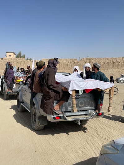 The image depicts an outdoor scene in Spin Boldak, Afghanistan, under a clear blue sky with bright daylight. The ground is unpaved, consisting of sand and dirt with visible tire tracks.
In the midground, a silver-grey Toyota pickup truck is positioned with its tailgate lowered. In the truck bed, a white sheet draped over a red fabric covers an elongated object, which rests on two carved wooden supports. Six men are visibly engaged with this object and the truck. Two men are seated on the lowered tailgate; one on the left wears brown traditional clothing and sandals, while the man on the right wears a green traditional tunic and sandals. Three men are standing in the truck bed, supporting the draped object; one wears a white turban, another a white turban and a blue garment, and a third a green cap and green garment. A sixth man, dressed in dark brown traditional attire and a dark head covering, stands in the truck bed facing forward.
Behind this truck, a white pickup truck is partially visible, also carrying multiple individuals standing in its bed, including a prominent figure in dark purple or blue fabric on the left. Objects resembling a light-colored frame and patterned fabric are also visible in the bed of the second truck.
In the background, a long, low structure made of light brown mud or adobe forms a rough wall across the scene. Behind this wall on the left, a small, two-story light-colored building with a flat roof is visible, along with sparse green foliage. A bicycle and other indistinct vehicles are present on the right side of the background, near the mud wall. A distant vertical structure, possibly a communication tower, is visible on the horizon above the mud walls.