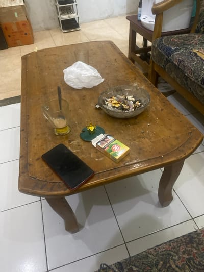 The image depicts an indoor scene within a living space in Cimenyan, Indonesia. A rectangular wooden coffee table, exhibiting surface wear and scratches, occupies the central foreground. On its surface, from left to right, are: a clear glass mug containing a small amount of light brown liquid and a silver spoon; a crumpled white plastic bag; and a clear glass ashtray filled with numerous cigarette butts, ash, and discarded foil. To the lower left of the ashtray, a red-backed smartphone with a dark, inactive screen lies face-up. Adjacent to the smartphone is a green cigarette pack, identifiable as "Gudang Garam 76," and behind it, a small green coaster topped with a golden crown-shaped decorative item. A small silver-colored rectangular object rests near the ashtray. The floor consists of large white tiles with dark grout lines. In the background, to the left, a brown cardboard box is visible against a light-colored wall. Towards the center-back, a white, multi-tiered shoe rack holds several pairs of casual footwear. To the right, part of a wooden armchair or sofa with patterned upholstery is visible, and behind it, a small wooden side table supports a white appliance, possibly a water dispenser. No individuals are present in the scene.