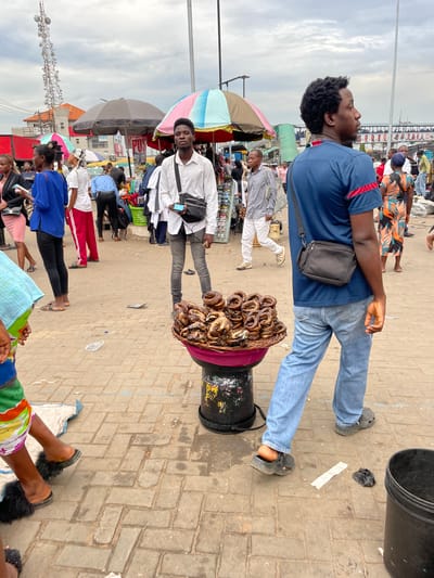 The image shows a busy market scene in Ikeja, Nigeria. Two men are prominent: one in a blue shirt and jeans, and another in a white shirt, both wearing shoulder bags. Between them is a black bucket, with a basket filled with what appears to be smoked fish balanced on top. The ground is brick-paved. Surrounding them are numerous people, market stalls with umbrellas, and various objects suggesting a vibrant, active marketplace. In the background, a communication tower can be seen.
