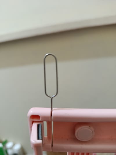 A close-up, eye-level shot shows a pink object with a silver, oblong loop sticking straight up from its top. The object appears to be made of plastic and has a textured surface. The background is out of focus, but it appears to be a wall with a horizontal line across it. The scene is indoors. The location is not specified in the image.