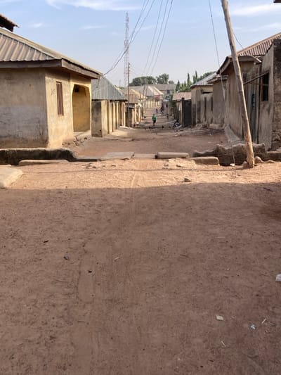 The image depicts a residential street in Minna, Nigeria, under clear daytime conditions. The foreground features an unpaved, reddish-brown dirt road with visible tire tracks and scattered debris. Two horizontal concrete slabs or blocks are positioned across the road in the immediate foreground. Single-story buildings with light-colored, weathered walls and corrugated metal roofs line both sides of the street, receding into the distance.

On the left, a house with a brown window frame and an arched entryway is partially visible. On the right, a wooden utility pole stands prominently, supporting overhead wires that extend down the street. In the mid-ground, a person wearing a green shirt walks away from the viewer, while other indistinct figures, including what appear to be children and small dark animals (possibly chickens), are visible further down the road.

The street slopes gently downward towards the background, where more residential structures are present. A tall, red and white striped lattice communications tower stands against the light blue sky in the distant background, with additional overhead wires visible beneath it. Shadows are distinct, consistent with direct sunlight.
