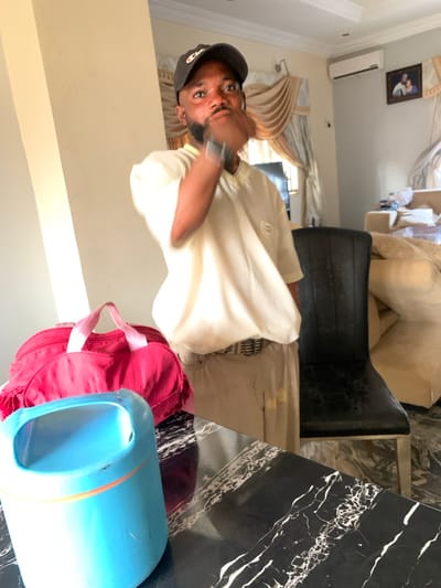 A person wearing a hat and a light-colored shirt is indoors. A black chair is near the person. A red bag and a blue container are in the lower right corner. In the background, there's a curtained window and a cream-colored sofa. A small framed picture is on the wall. The image appears to be rotated.