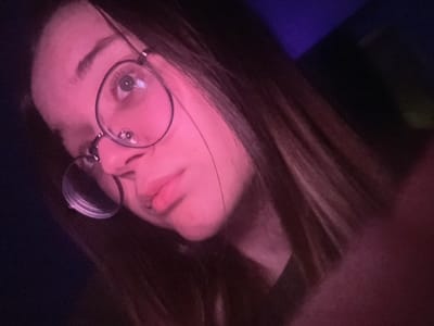 A close-up, low-angle portrait features an individual, likely female, with long dark hair and pale skin, illuminated primarily by a strong pink/magenta light source from the left. The subject is wearing round, clear-rimmed glasses with dark metal frames, through which their right eye (viewer's left) is visible looking upwards and slightly to the right. The mouth is closed with a slight pout. The pink light heavily colors the subject's face, neck, and portions of the hair. The background is dark and indistinct, with a muted purple area visible in the upper right quadrant. The specific location within the city of Pushchino, Russia, cannot be determined from the visual information presented in this image.
