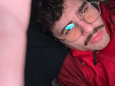 A person with dark curly hair and a prominent mustache is reclining. They are wearing round gold-rimmed glasses and a red zippered jacket. The person appears to be lying on a black surface, possibly a couch or bed. A blurry pink shape is visible on the left side of the frame, possibly an arm or leg close to the camera. The individual's expression is slightly quizzical or skeptical.