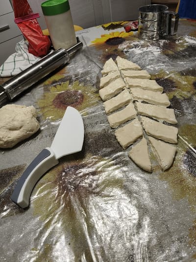 This image captures a close-up, elevated view of a kitchen table in Villa di Gemmano, Italy, during a food preparation activity. The table is covered with a clear plastic tablecloth featuring a yellow sunflower pattern against a darker background, and is dusted with white flour.
In the upper left, a silver-colored metal rolling pin rests horizontally on a white and green checkered cloth. Behind it, a tall, cylindrical white container with a bright green lid stands upright, next to a partially visible red package secured with a pink clip. In the upper right background, a silver-colored metal flour sifter is present.
The primary subjects are various forms of raw, light beige dough. A rounded mound of dough is positioned on the mid-left. To its right, a series of approximately 20-25 irregular rectangular and triangular dough pieces are arranged in a branching pattern, indicating they have been recently cut from a larger mass. Between the dough mound and the cut pieces lies a white and gray dough scraper with a curved blade and a gray ergonomic handle. Scattered flour particles are visible across the entire surface of the tablecloth, particularly around the dough and utensils. No individuals are present in the scene.