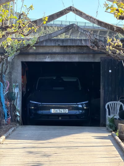 The image shows a dark blue car parked inside a garage. The car's license plate, "BX 74 TD," is visible. The garage is located between a concrete wall and a stone wall. The entrance to the garage is partially obscured by branches and foliage. In the area around the garage entrance, there are various items including a white plastic chair, debris, and some ropes. The setting appears to be in Vila Real, Portugal, based on the license plate format, which indicates a Portuguese vehicle registration.