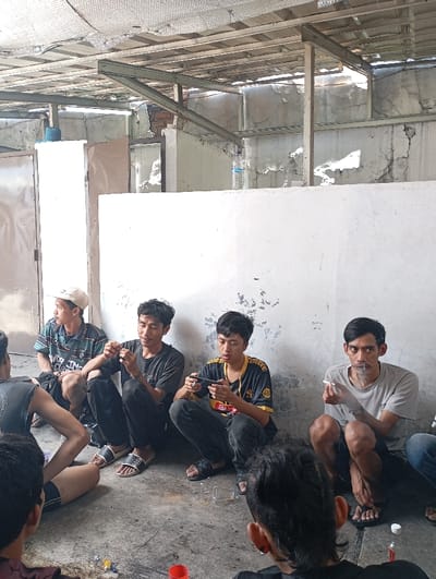 The image shows five men sitting on the floor in a partially constructed building. The location is Katapang, Indonesia, at latitude -6.988, longitude 107.557. Three of the men are sitting facing the viewer, while one sits with his back to the camera and the fifth man is out of frame. The men are all dressed casually. One man is holding a mobile phone, while another is holding a cigarette. The floor is concrete and there are visible signs of unfinished construction.