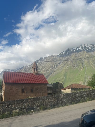 A stone church with a red metal roof and a bell tower sits in front of a mountain in Stepantsminda, Georgia. The church has two small windows and is surrounded by a stone wall. A second, smaller stone building with a terracotta tile roof is visible to the right of the church. In the background, a large mountain with snow-capped peaks rises steeply, its slopes covered in green grass. The sky above is a mix of blue and white clouds. Power lines stretch across the frame in front of the mountain. A paved road is in the foreground, and the front of a black car is visible in the lower right corner.