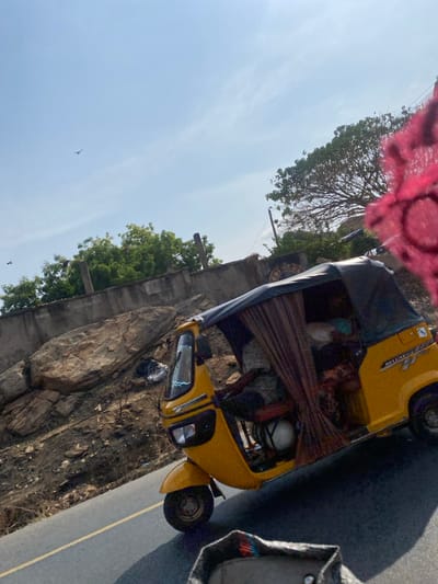 This image captures a daytime scene in Ogaminan, Nigeria, depicting a bright yellow three-wheeled motorized rickshaw (keke napep) on a paved road. The rickshaw, labeled "DELUXE 177" and "CNG," features a black fabric canopy and brown patterned curtains partially covering its passenger compartment. A driver is discernible within, obscured by the curtains, and additional forms (possibly passengers or cargo) occupy the rear. A white, helmet-like object rests on the floor of the driver's side.
The road features a double yellow line. To the immediate left, a steep, rocky embankment dominates the midground, characterized by large, irregular brown boulders and sparse, dry ground vegetation, with some areas appearing burnt. Above the embankment, a weathered concrete wall with vertical posts separates the rocky terrain from lush green foliage and trees in the background. A large tree with extensive branching is prominent on the right side of the frame, with a utility pole nearby. The sky is clear and light blue, with a single bird visible in flight in the upper left quadrant. A partial circular sign, dark with light lettering (possibly "COSMAS"), is affixed to the concrete wall above the rickshaw. A portion of a dark, textured object with red, orange, and blue stripes is visible in the bottom foreground, suggesting the vantage point of the photographer. A bright pink, patterned fabric is partially visible in the extreme upper right corner.