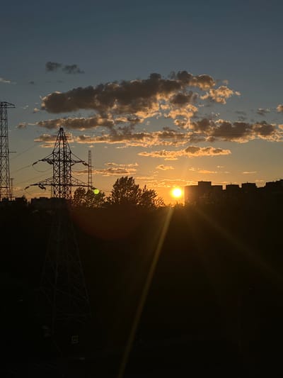 The image shows a sunset over the city of Shushary, Russia. The sun is low on the horizon, casting a warm golden light on the clouds and silhouetting the surrounding landscape. In the foreground, a large electrical pylon stands tall, its metal structure stark against the colorful sky. Trees and buildings are visible in the middle ground, also silhouetted by the setting sun. The sky is a gradient of blues and oranges, with scattered clouds tinged with yellow. The overall impression is one of a peaceful evening in an urban environment.