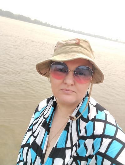 A medium shot captures a woman from the chest up, standing on a body of water with a shoreline visible in the distance. She is wearing a bucket hat and large, rimless sunglasses with a pink gradient. Her top has a bold, geometric pattern of black, white, and blue shapes. A thin gold necklace with a cross pendant is visible. The water is calm and appears slightly murky, with a hazy sky and a tree line along the distant shore. The scene is set in Bibikha, Russia, a locale characterized by its proximity to water and natural landscapes.