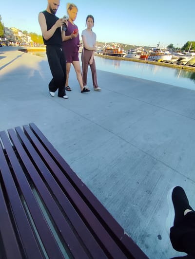 In Sarıyer, Turkey, three individuals walk along a concrete promenade bordering a marina. The person in the center, a man with a beard, wears a sleeveless black shirt and black pants, holding a smartphone. To his left, a woman in a burgundy top and shorts looks towards the man, also holding a smartphone. To the man's right, a woman in a white t-shirt and brown pants walks with her head down. The promenade has a dark wooden bench in the foreground, with its slats angled away from the viewer. In the background, numerous yachts are docked in the calm water of the marina, with buildings and greenery visible on the distant shore under a clear blue sky.