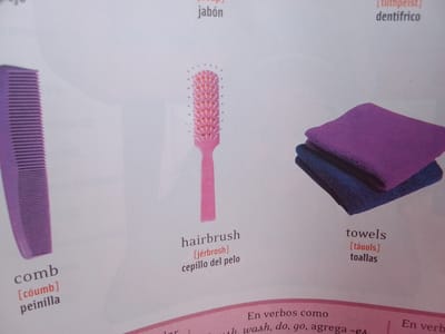 This media file is a photograph of a page from a vocabulary book. The page displays illustrations and text related to personal hygiene and household items.

The subjects are objects: a purple comb, a pink hairbrush, and a stack of two towels (one purple and one dark blue). The objects are presented in a clean, studio-like setting against a plain white background, which is typical for educational materials.

The activity taking place is the presentation of vocabulary words. The page is divided into sections, each featuring an image, its English name, a phonetic pronunciation guide, and its Spanish translation.

Notable details include the text present on the page. For the comb, the text reads "comb [cóumb] peinilla". For the hairbrush, it reads "hairbrush [jérbrosh] cepillo del pelo". For the towels, it reads "towels [táuols] toallas". There is also fragmented text visible at the bottom of the page, suggesting a section on verbs: "En verbos como whwash, do, go, agrega -e En verb".

The time of day, weather, location cues (other than it being a vocabulary book page), and emotions are not discernible from the image. The overall presentation is straightforward and educational. The focus is clearly on the vocabulary being taught, with no extraneous elements. The quality of the image suggests it was taken indoors, likely with artificial lighting.