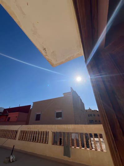 The low-angle shot looks upward from a courtyard, perhaps in the city of Ouaouizert, Morocco. The deep blue sky is pierced by the sun's bright rays. The buildings surround the scene, exhibiting traditional architecture in earth tones, with some decay visible. A building on the right shows clothes hanging out a window. A wooden structure is visible at the top left.