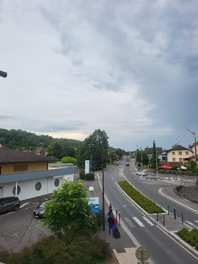 The image shows a street scene in Publier, France, taken from a high vantage point. The scene is dominated by a gray, overcast sky with patches of blue. The road, which has white lane markers, is empty of traffic. There are two figures walking on the right side of the road, one in blue and one in gray. To the left of the road is a parking lot with two cars. To the right of the road are green bushes and trees. There is a building with a red roof in the distance, and another building with a yellow facade to the left of the parking lot. A sign with blue and white lettering is visible on the right side of the road. A manhole cover is visible in the foreground.