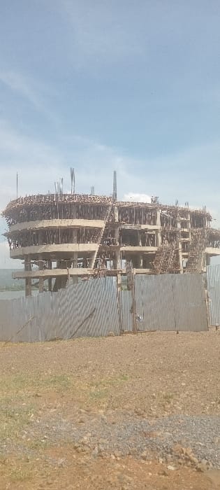 The image displays a multi-story concrete structure under active construction, situated along the waterfront in Kisumu, Kenya, likely bordering Lake Victoria. The building exhibits a circular or oval footprint with at least four discernible levels of concrete framework, extensively supported by wooden scaffolding and formwork. Steel reinforcing bars protrude from the uppermost sections, indicating ongoing vertical development. Three indistinct human figures are observed on the top level of the structure, presumably engaged in construction activities. The foreground consists of dry, reddish-brown soil and gravel with sparse vegetation, delineated by a corrugated metal fence approximately 1.5 to 2 meters in height. A large body of water is visible in the distant background under a light blue sky with scattered white clouds.