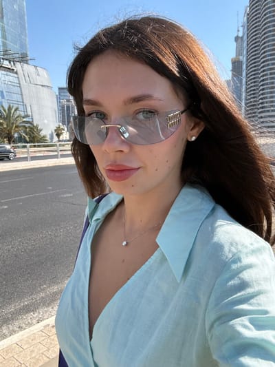 A medium shot shows a young Caucasian woman with long, dark brown hair and fair skin, wearing large, futuristic-style sunglasses with a light blue tint and gold accents. She is wearing a light blue collared shirt and a delicate silver necklace with a single diamond pendant. A small, diamond stud earring is visible in her right ear. The background features a city street scene with palm trees, modern buildings, and a bright blue sky. The woman is looking directly at the camera. The location is identified as the city of Al Jizah, Jordan.