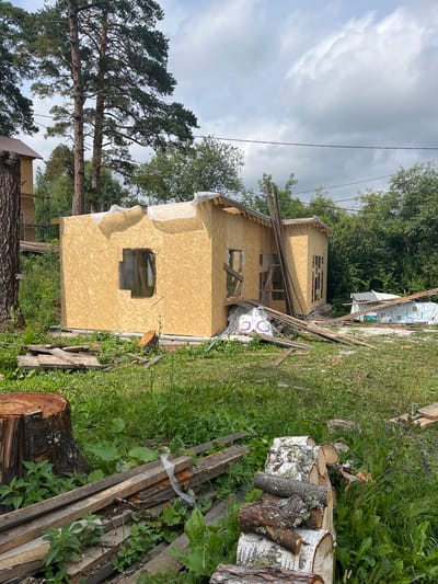 The image shows a partially constructed wooden house with walls made of oriented strand board (OSB). The structure is in an outdoor setting with tall trees and green foliage. Several pieces of lumber are scattered around the site, along with some construction materials wrapped in plastic. A large tree stump is visible in the foreground. The sky is overcast with clouds. The scene appears to be a rural or suburban construction site.