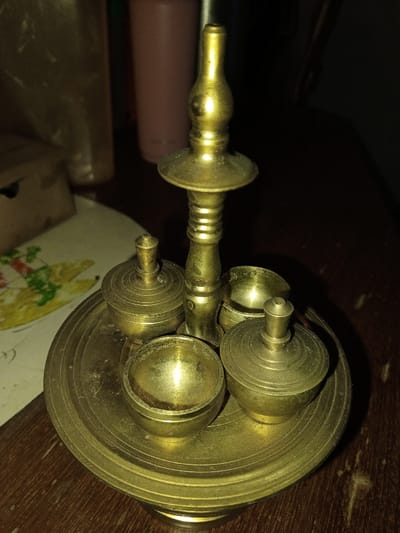 The image displays a multi-component brass artifact, resting on a dark brown wooden surface. The artifact consists of a central, ornate column with a tapered, spired finial at its apex, rising from a circular, tiered base. The base features concentric etched lines on its flat surface. Arranged symmetrically on this circular base are four smaller brass vessels: two are lidded, dome-shaped containers with small finials on their tops, and two are open, hemispherical bowls. All brass components show signs of tarnish and some surface accumulation. In the blurred background, a light-colored paper with a colorful, possibly floral pattern (red, green, yellow) is visible on the left. Further back, a tall pink cylindrical container and a translucent plastic bag are present. No specific visual cues within the image confirm the location as Longmiang, Laos.