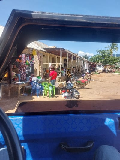 The image depicts an outdoor marketplace scene in Kimamba, Tanzania, viewed from the interior of a blue vehicle, with the upper part of the vehicle's window frame visible at the top and the patterned blue door panel, along with a handle and a partial view of a leg in blue jeans, at the bottom. The sky is clear blue with minimal white clouds.
The ground surface is unpaved dirt. On the left, a vendor stall is set up, featuring various items of merchandise, including colorful fabrics and other goods, hanging from a wooden structure. A person in a patterned dress is standing near this stall, and another individual is partially visible behind a wooden table with items on it. Three plastic chairs, two blue and one green, are positioned around a small wooden table, where at least two individuals appear to be seated or conversing. A person in a red shirt stands nearby.
Further down the unpaved path, a row of single-story buildings with corrugated metal roofs extends, supported by wooden posts forming a covered walkway or veranda. These buildings appear to house additional market stalls or businesses. Multiple motorcycles are parked on the dirt path; one black motorcycle with a visible Tanzanian license plate "T593 DRE" is centrally positioned. Another motorcycle, partially visible further back, is green and red. Various other objects, including buckets and containers, are scattered around the scene. A large green tree and a palm tree are visible in the background on the right.