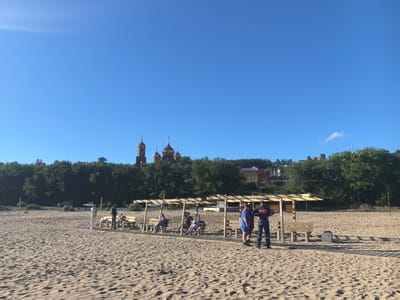 The image depicts a sandy beach under a clear blue sky, with a line of trees and buildings in the background. In the distance, several golden-domed buildings, likely churches, are visible. On the beach, there are several wooden structures providing shade, with benches beneath them. Some people are seated on the benches, while others are standing or walking on the sand. Two men in what appear to be uniforms are standing near one of the shaded areas. There are also some cars parked along a road bordering the treeline. The scene suggests a public recreational area on a sunny day. The architecture of the domes and the presence of buildings on a hill are characteristic of the Znamensky Monastery in Tolyatti, Russia, which is located on the banks of the Kuybyshev Reservoir.