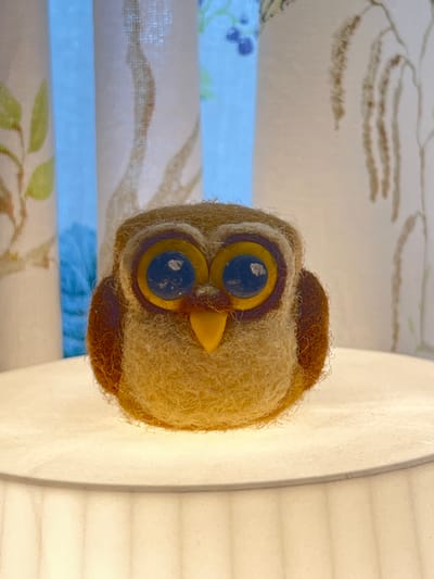 A needle-felted owl figurine sits on a lampshade. The owl is predominantly light brown with darker brown accents around its eyes and wings. It has large blue eyes with yellow and purple rings around them and a yellow beak. The owl is positioned centrally on a white, circular lampshade, which is illuminated from below, casting a soft glow upwards onto the figurine. In the background, sheer curtains with a subtle floral pattern are visible. The curtain on the left has a light blue hue showing through it, suggesting daylight or ambient light coming from outside. The scene is indoors. There are no people or interactions depicted.