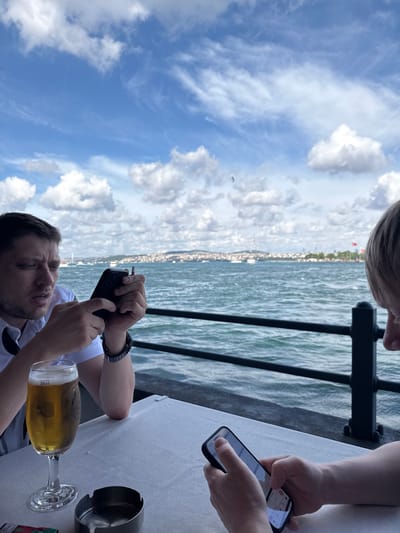 The image depicts two individuals seated at a table by the water in Istanbul, Turkey. On the left, a man is holding a smartphone in his right hand and a cigarette in his left, appearing to be engaged with the device. A glass of beer is positioned on the table to his left. The man is wearing a white collared shirt. To the right, another person, whose face is not fully visible, is also holding a smartphone and appears to be interacting with it. The table is covered with a white tablecloth, and an ashtray sits on the table between the two individuals. In the background, the Bosphorus Strait is visible, with boats and the cityscape of Istanbul stretching across the horizon under a blue sky with scattered clouds. A black metal railing separates the seating area from the water.