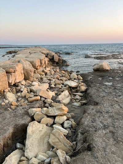The image displays a rocky shoreline with the sea on the left and a pile of stones in the foreground. The stones vary in size and color, ranging from light beige to dark gray. Some of the stones are large and irregular, while others are smaller and more angular. The sea is a deep blue-gray and appears to be calm, with a slight ripple on the surface. The sky is overcast, with a faint rainbow visible in the upper left quadrant of the image. The scene appears to be set in a natural, outdoor environment. There are no people or animals visible in the image. The overall mood of the image is one of tranquility and natural beauty. The lighting suggests it is either early morning or late afternoon. The location appears to be a coastal area. There is no visible text in the image.
