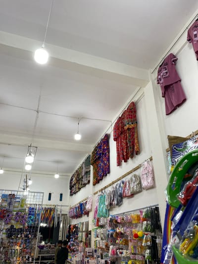 The image shows the interior of a brightly lit store, likely a market or a shop selling various goods. The perspective is from a low angle, looking upwards towards the ceiling and shelves. The scene is densely packed with merchandise. On shelves along the walls, folded textiles, possibly clothing or fabric, are neatly stacked. These items display a variety of patterns, including plaid and floral designs, in vibrant colors like red, yellow, purple, and green.
Above these, on what appears to be a higher shelf, are more packaged items, possibly accessories or toys, with some featuring bright colors and cartoonish designs. In the foreground and extending into the background, metal racks are filled with a wide assortment of products. These include colorful umbrellas, sunglasses with playful designs, small stuffed animals, keychains, and other trinkets.
The store's lighting is provided by several round, white light fixtures attached to the ceiling and walls, creating a well-illuminated environment. In the background, a person wearing a dark jacket is visible, seemingly browsing or working in the store. The overall impression is that of a busy retail space offering a diverse range of affordable items. The context of "Lamteumen Barat, Indonesia" suggests this is a local shop in that region. There is no clear indication of the time of day or weather conditions, as the interior lighting is the dominant light source. Some text is visible on product packaging, but it is too small and blurry to read clearly.
