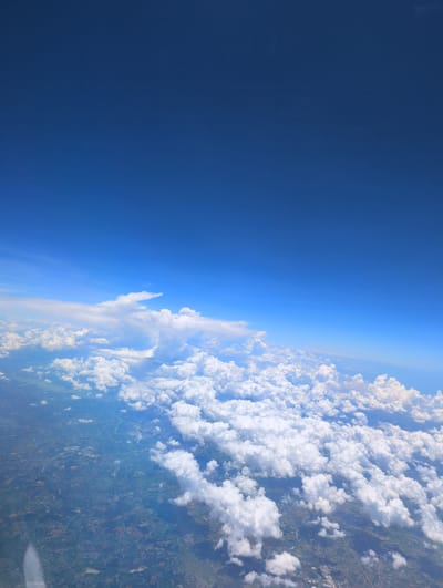 The image is an aerial view of a vast expanse of sky and clouds, likely taken from an airplane. The upper portion of the image is dominated by a clear, deep blue sky. Below this, a band of fluffy white clouds stretches across the horizon. These clouds vary in density and shape, with some appearing wispy and others more voluminous.

Beneath the clouds, the earth is visible, appearing as a patchwork of green and brown terrain. Roads and structures, indicating a populated area, can be discerned, though they are small and indistinct from this altitude. The overall impression is one of great height and distance, with the land below appearing miniature. The specific location in Zhanjiang City, China, cannot be determined from the visual information provided in the image.