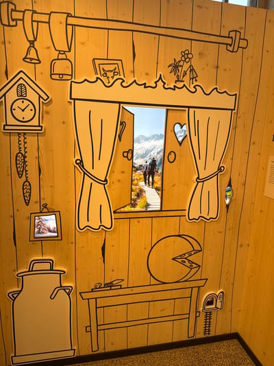 The image depicts a decorative wooden wall in the city of Schattendorf, Switzerland, designed to resemble the interior of a rustic dwelling with cartoon-like illustrations. The wall is adorned with black outlines of various objects, including a cuckoo clock, bells, a milk can, a table with a cheese and knife, and a small mouse door with a miniature railroad track. A prominent feature is a large, stylized window frame with drawn curtains, through which a photograph of two hikers on a wooden boardwalk in a mountainous landscape is visible. The overall scene conveys a sense of alpine charm and a whimsical, storybook aesthetic.