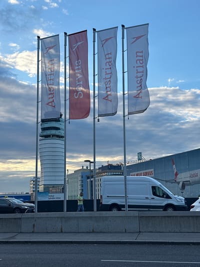 The image shows a street view in Schwechat, Austria, displaying a row of Austrian Airlines flags fluttering in the wind. The flags feature the airline's logo and the word "Austrian," with one flag displaying "Servus." Behind the flags, the architectural elements of Vienna Airport, including the air traffic control tower, are visible. A white van with the inscription "DAC NEUE SERVUS" is parked along the street, and several cars are parked behind it. A person wearing a yellow shirt is seen in the background. The sky is partially cloudy.