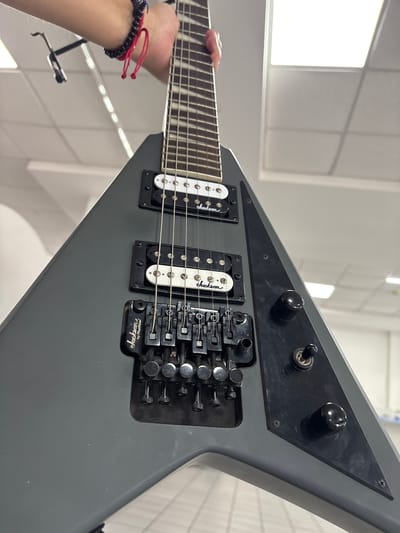 The image shows a close-up, low-angle shot of a dark gray Jackson V-shaped electric guitar, held by a person whose arm and hand are visible at the top left. The hand wears a dark beaded bracelet with red string accents. The guitar's fretboard, two humbucker pickups with white and black covers (both labeled "Jackson"), and the Floyd Rose locking tremolo system are clearly visible. The guitar has a matte finish. The background appears to be an indoor setting with ceiling lights and what looks like office or retail shelving. There is no discernible indication of the specific location in Cuernavaca, Mexico, as the image focuses entirely on the guitar and the person holding it.