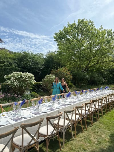 The image shows an outdoor scene with a long banquet table set for a meal. The table is covered with a white tablecloth and has place settings consisting of plates, silverware, glasses, and menus. Blue flowers are placed in vases down the center of the table. There are wooden chairs with off-white cushions set on both sides of the table. Two women are standing near the table, appearing to be interacting or posing for a photo. The background includes green trees, shrubs, and grass, suggesting a garden or park setting. The scene evokes a sense of an outdoor event or celebration. The location is near Cambridge, United Kingdom.