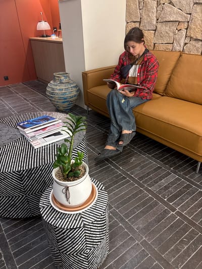 A young woman with brown hair is sitting on a tan leather sofa reading a book. She is wearing a red and black plaid shirt over a brown t-shirt, blue jeans, and dark grey slip-on sandals. A black fanny pack is worn across her chest. The sofa is against a wall covered with natural stone. In front of the sofa is a patterned ottoman with black and white geometric designs, stacked with magazines. To the right of the ottoman is a smaller patterned stool with a white potted plant. Behind the ottoman and stool is a large blue and white patterned vase. The floor is tiled with dark grey rectangular tiles laid in a herringbone pattern. The environment appears to be an indoor space, possibly a waiting area or lounge, with warm lighting. The image does not provide any specific indicators for the location in Busan, South Korea.