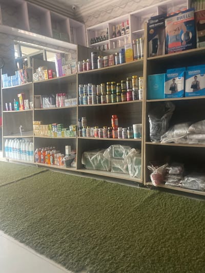 The image depicts a pharmacy or a retail store stocking various health and personal care products. The store appears to be located in Lafia, Nigeria. Shelves are filled with a diverse range of items, including bottled liquids, boxes of supplements, personal hygiene products, and medical supplies. The products are arranged in an organized manner, with labels and packaging visible for many items. The store's interior is simple, with plain shelving units and a green carpeted floor in the foreground. The lighting suggests an indoor setting, possibly with natural light coming from a window or doorway. The overall impression is of a well-stocked local business.